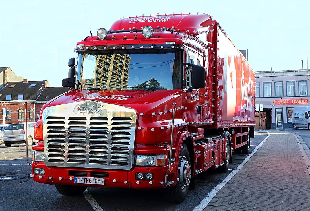 Un camion de Noël Coca-Cola rouge décoré de lumières, stationné sur une place.
