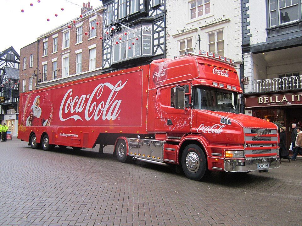 Un camion de Noël Coca-Cola illuminé, stationné dans une rue commerçante le soir.