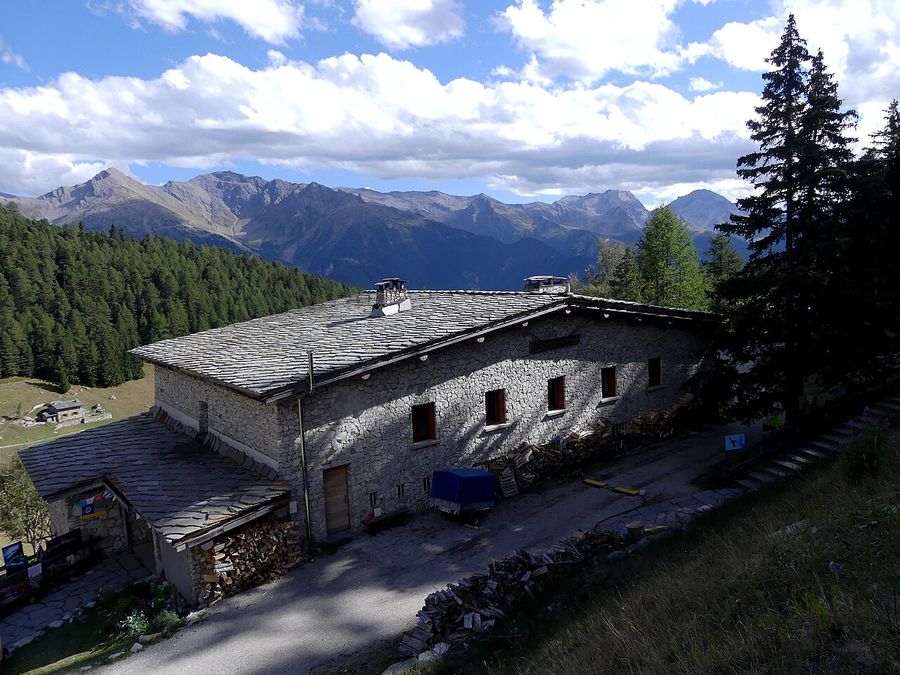 Refuge de montagne en pierre au toit de lauzes, avec des montagnes et des nuages en arrière-plan.