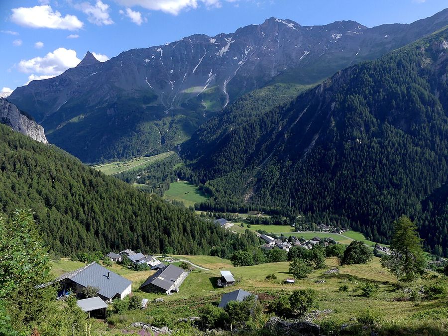 Vallée alpine verdoyante avec un hameau aux toits gris, des forêts et des sommets enneigés.